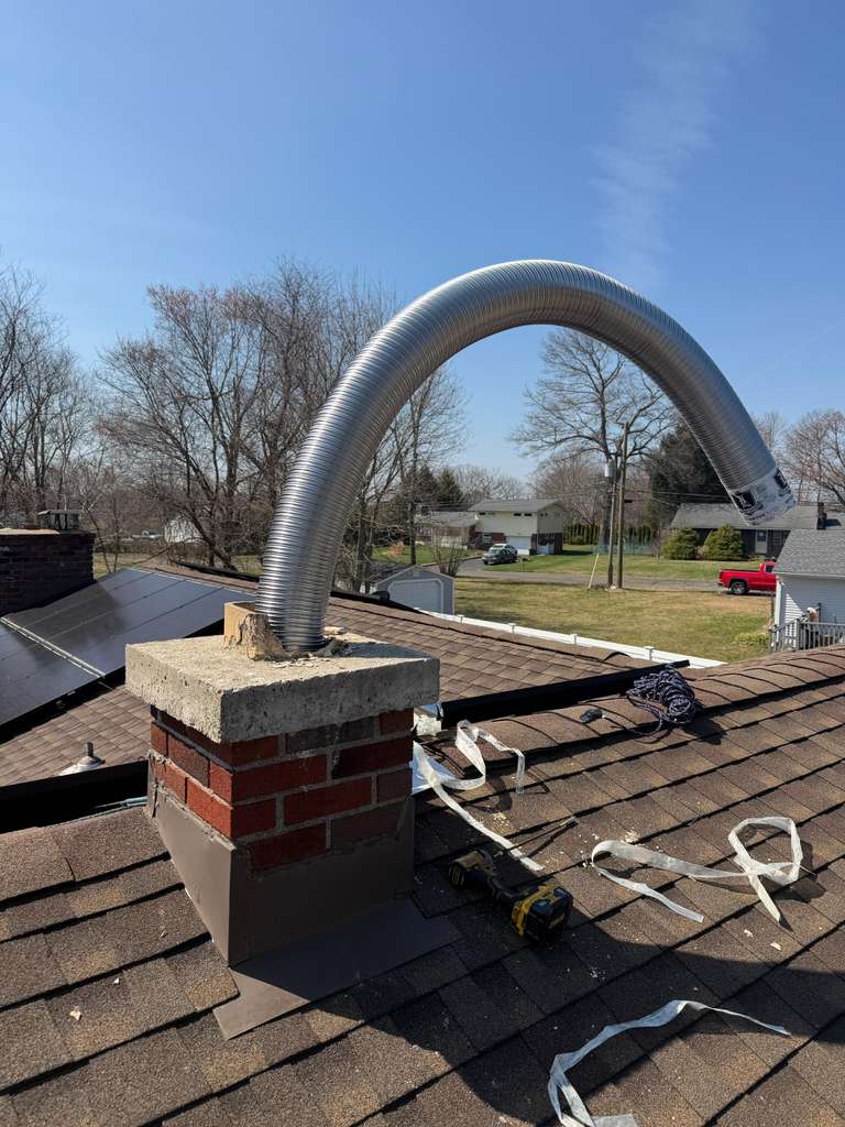 Flexible metal chimney liner arching from a brick chimney on a shingled residential roof.