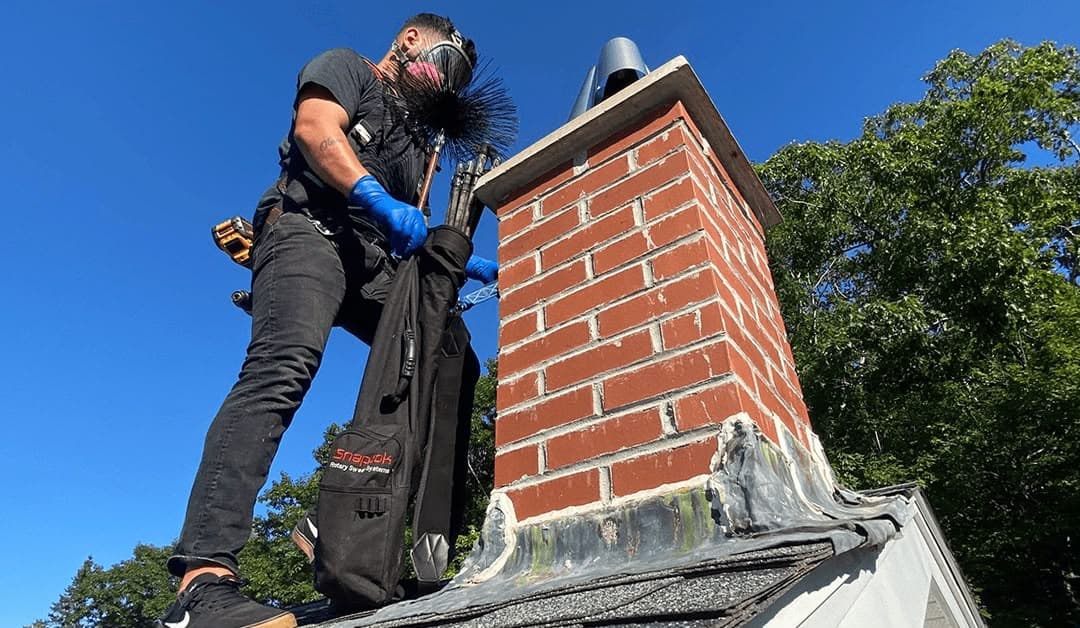 Professional chimney sweep with tools stands on a roof next to a red brick chimney.