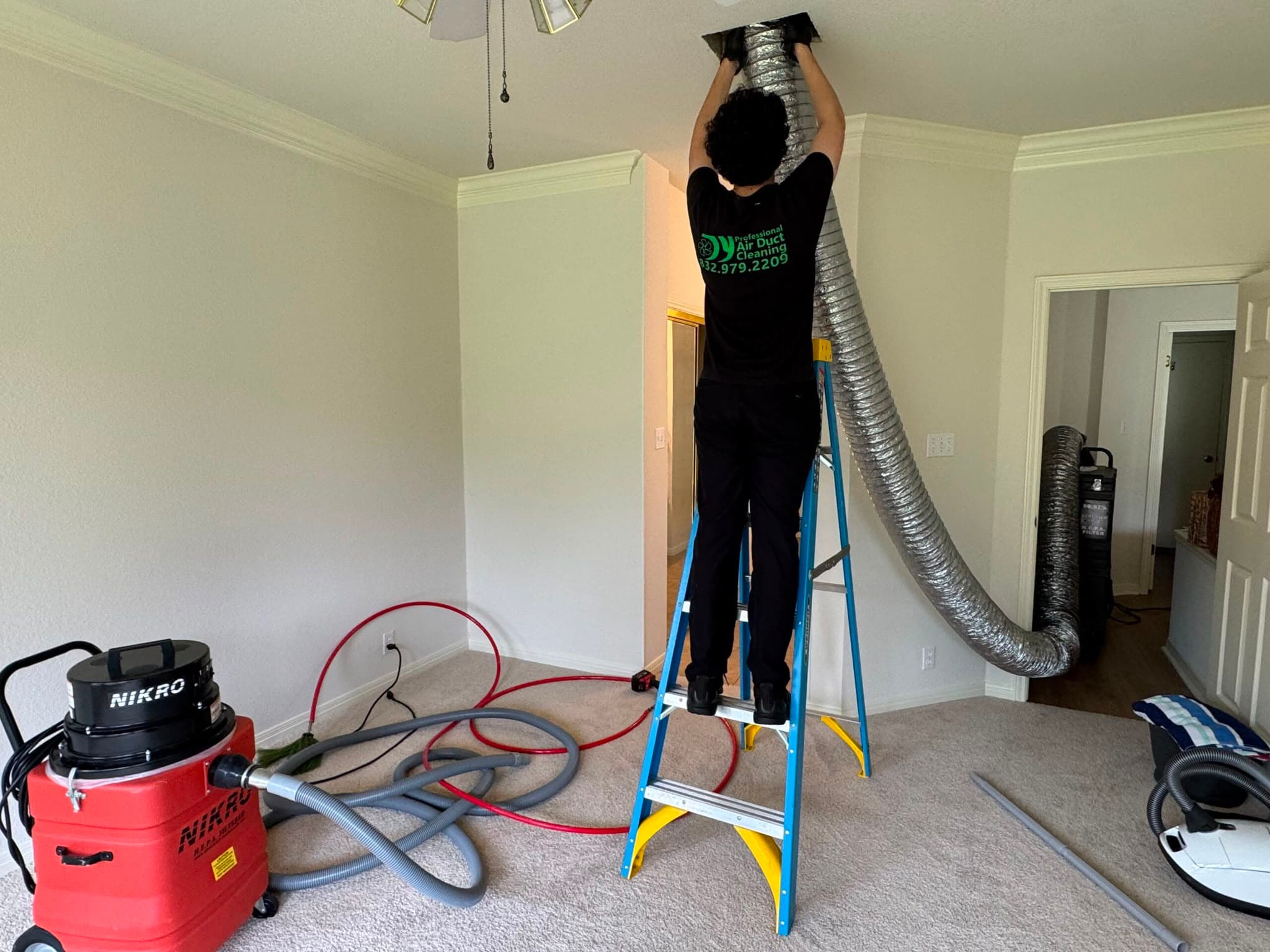 Technician on a ladder cleans ceiling air ducts with a large hose and industrial vacuum.