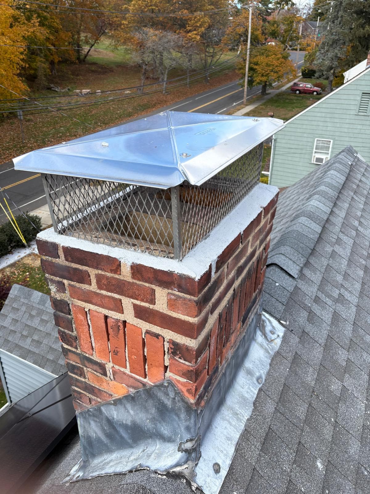 Red brick chimney with a metal cap and mesh screen on a grey shingled roof.