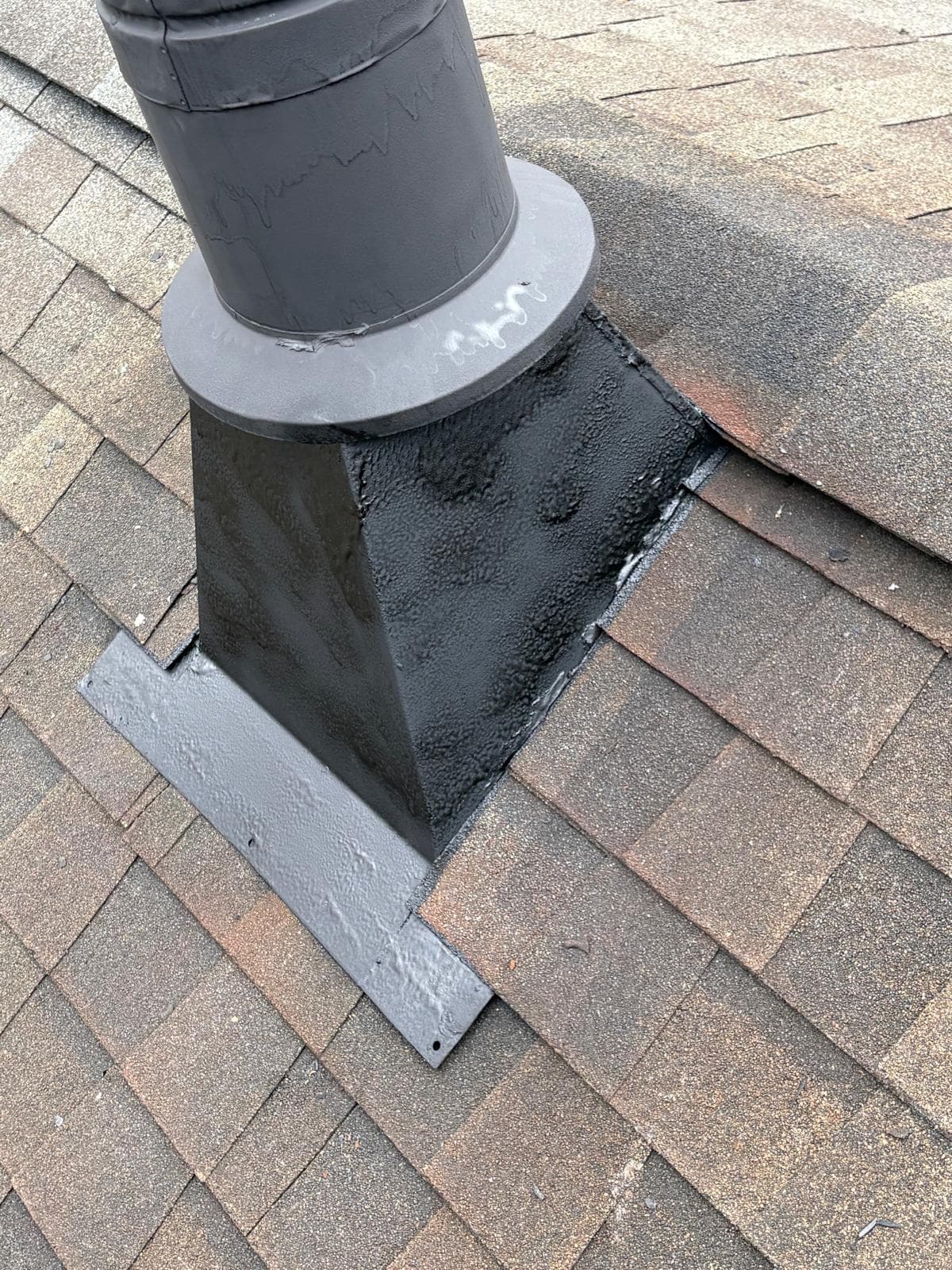Black metal roof vent pipe sealed with thick textured sealant on brown asphalt shingles.