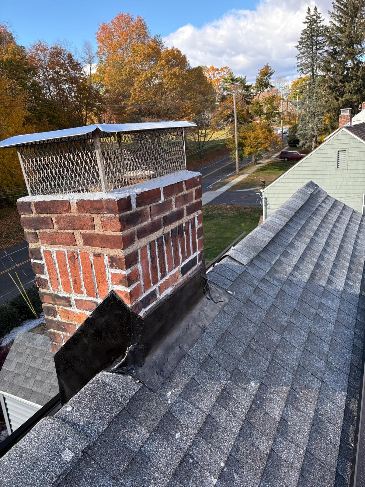 Brick chimney with a metal cap on a shingled roof overlooking neighborhood autumn foliage.