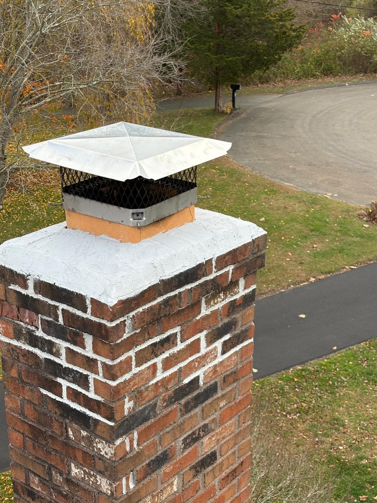 Red brick chimney with a metal cap and white sealant overlooking a grassy yard.