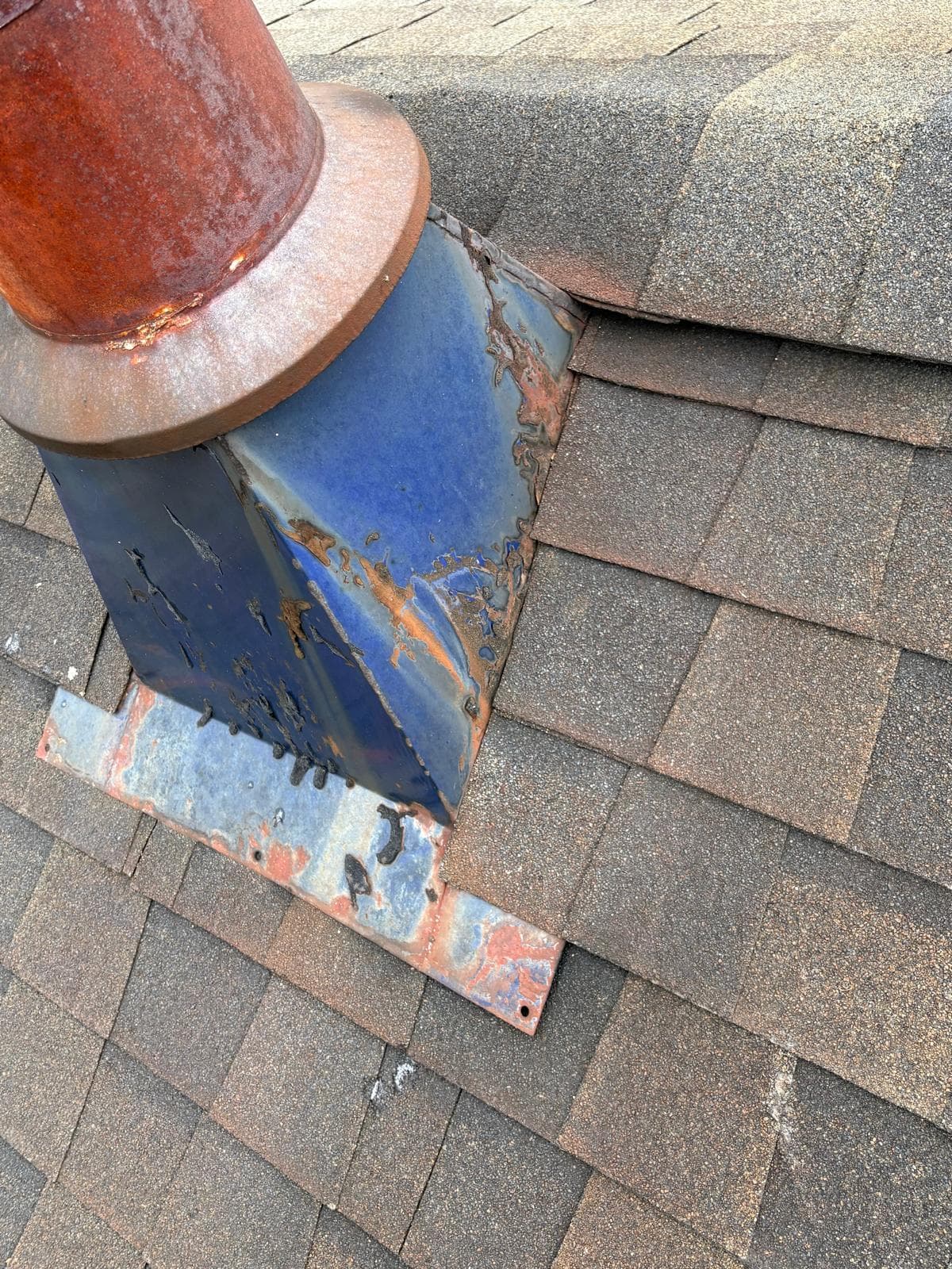 Rusted blue metal chimney vent and flashing showing severe corrosion on a shingled roof.