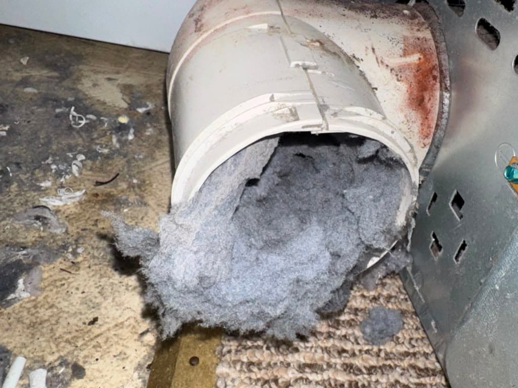 A white plastic dryer vent pipe heavily clogged with a thick mass of grey lint.