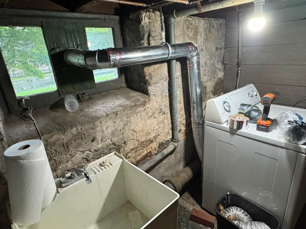 Basement laundry room with washing machine, utility sink, and metal vent pipe exiting a window.