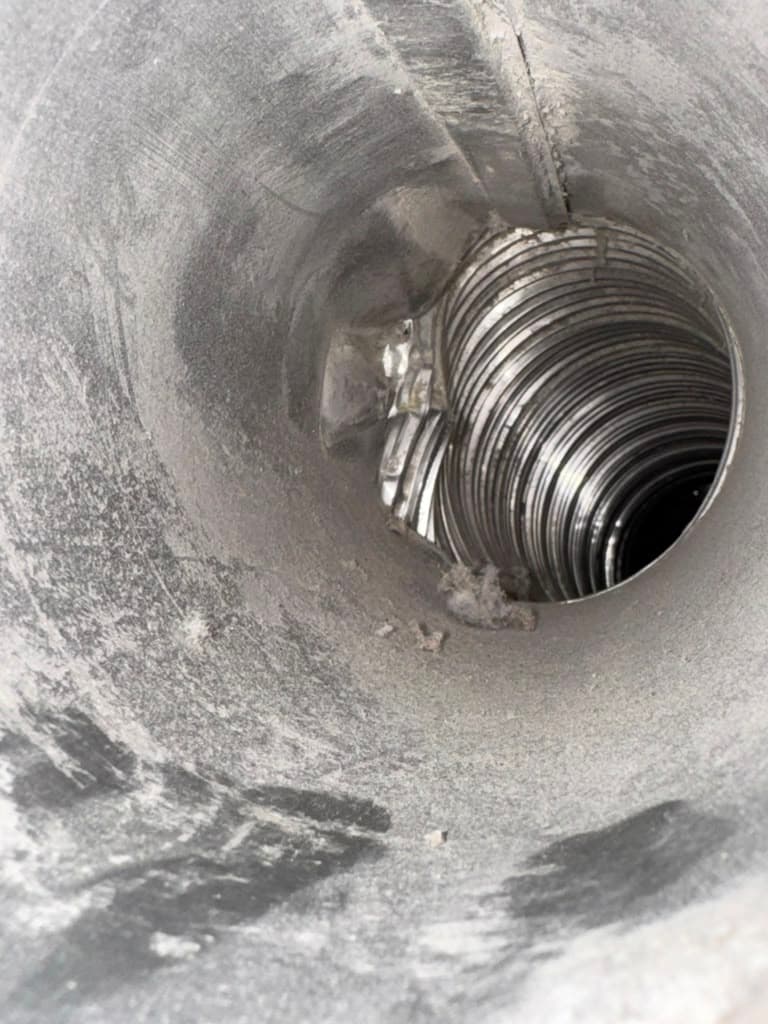 Interior view of a dusty metal air duct connecting to a flexible, ribbed silver ventilation pipe.