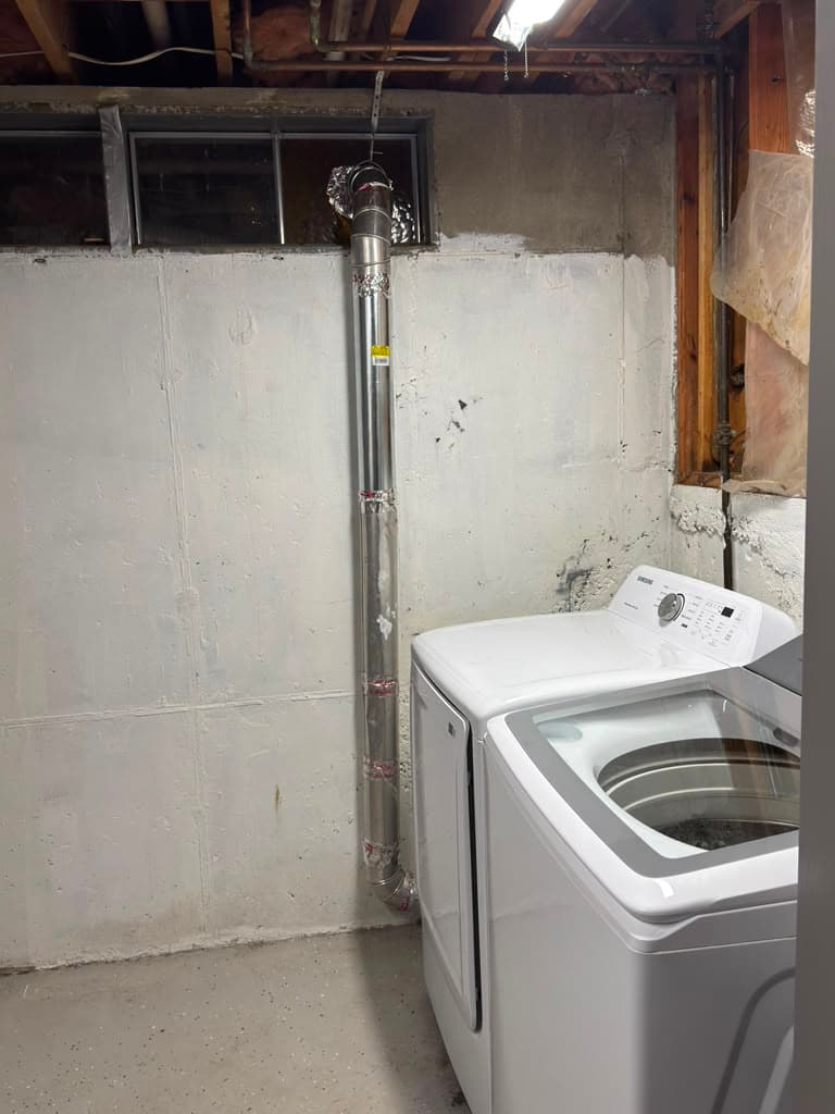White laundry appliances in a basement with a tall vertical silver metal exhaust vent.