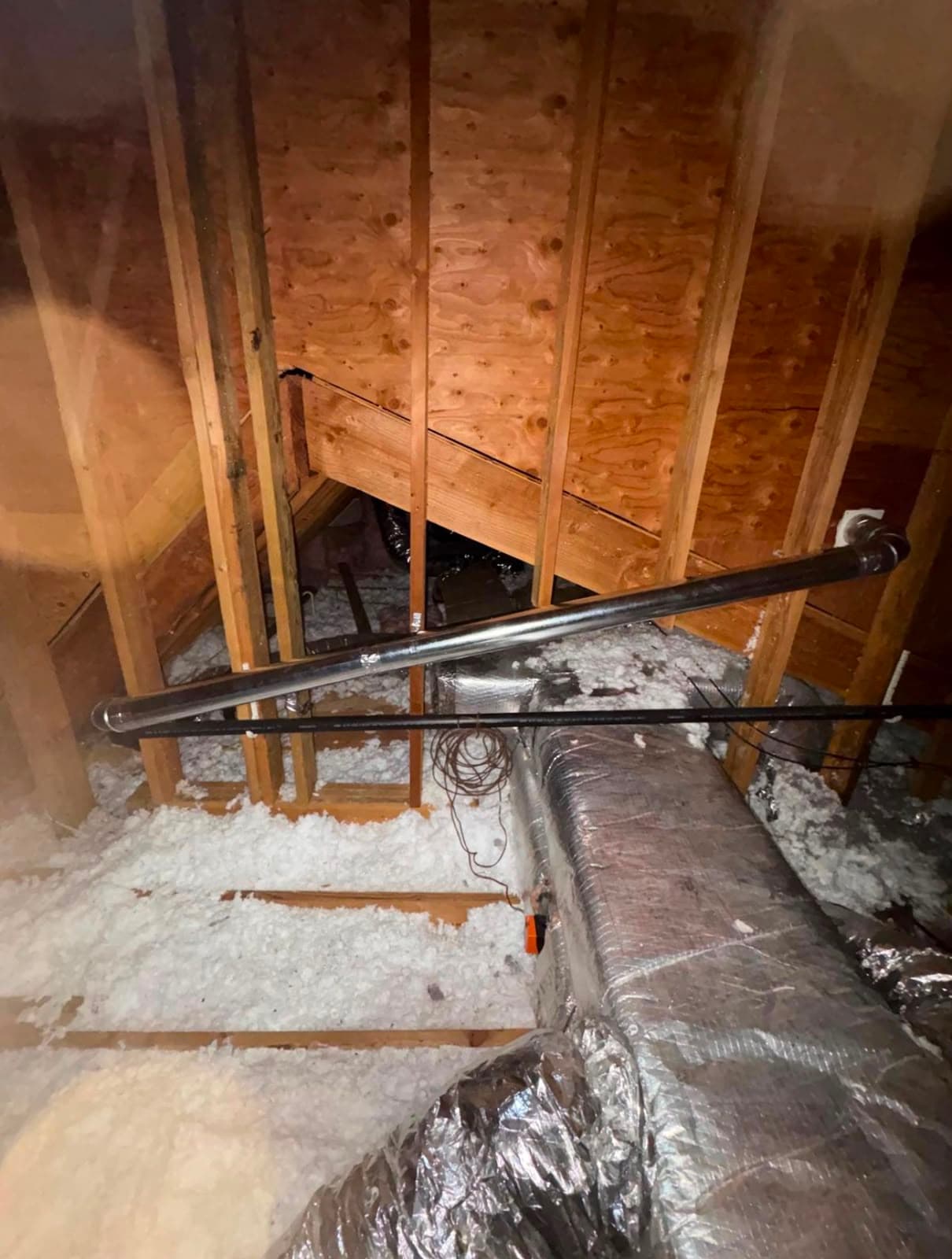 Attic interior featuring wooden framing, white insulation, silver HVAC ducts, and metal piping.