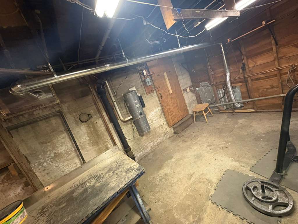Dimly lit basement with concrete walls, exposed ceiling pipes, a wooden workbench, and gym equipment.