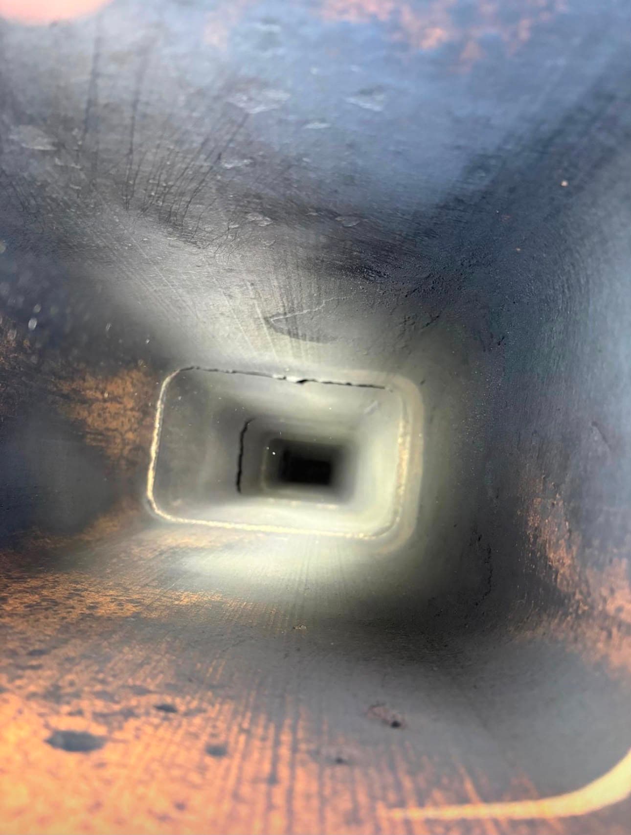 View down a dark, rectangular chimney flue with soot-covered walls and light at the end.